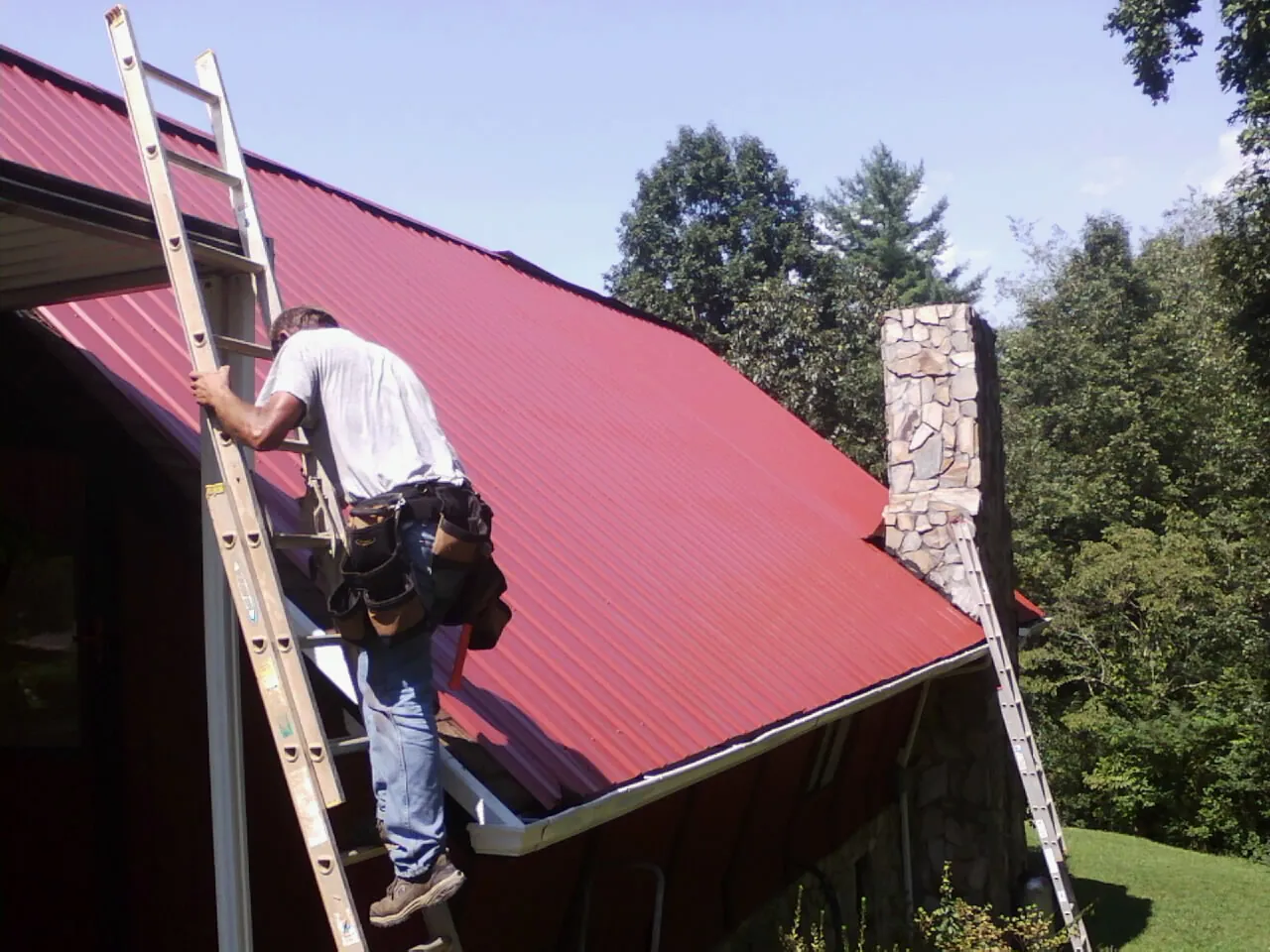 Roofer on a ladder beside a red metal roof and stone chimney — Soffit & Fascia Repair by Roofing Brothers in Carlsbad